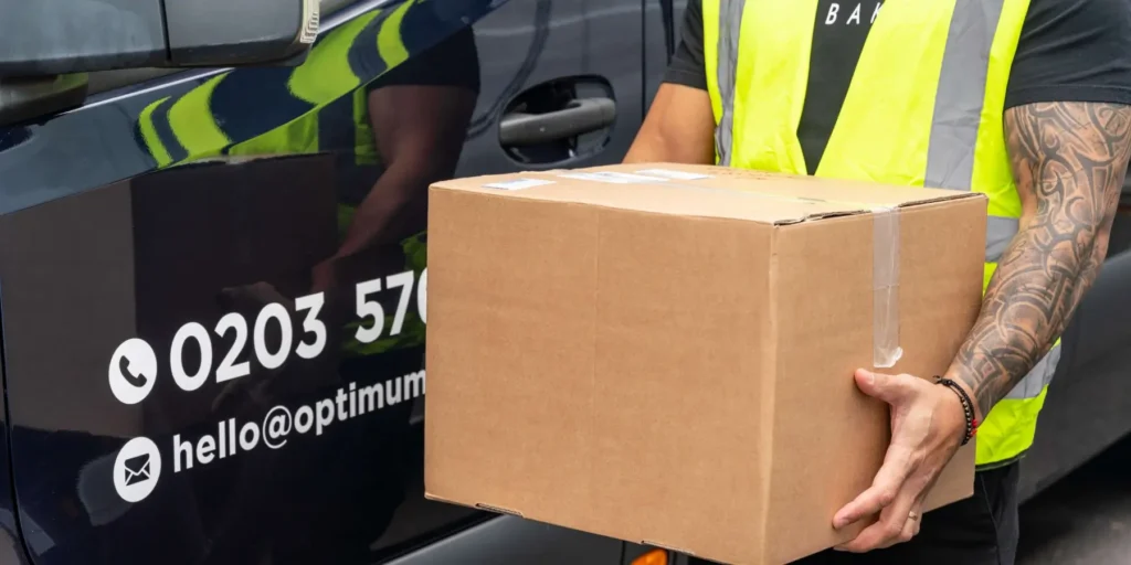 ourier in high-visibility vest carrying a cardboard box next to a dark blue branded delivery van with contact details on the side.